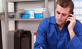 A young electrician or technician in a blue work shirt talks on a phone while sitting in what appears to be a service van or work vehicle with storage compartments visible in the background.